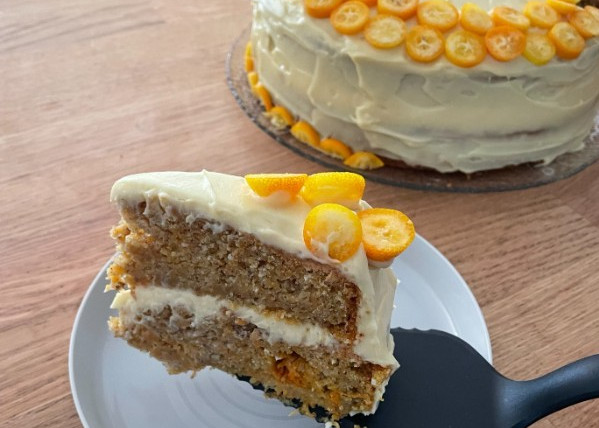 Bild eines Stücks Möhren-Orangen-Torte auf einem Tortenheber über einem kleinen Teller. Den Rest der Torte ist im Hintergrund zu sehen.